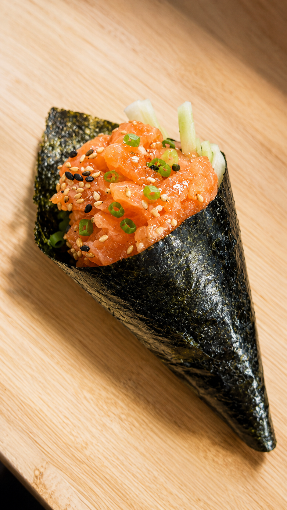 A single cone-shaped sushi wrap filled with orange salmon, white rice, green cucumber, and topped with sesame seeds, placed on a light wood surface.