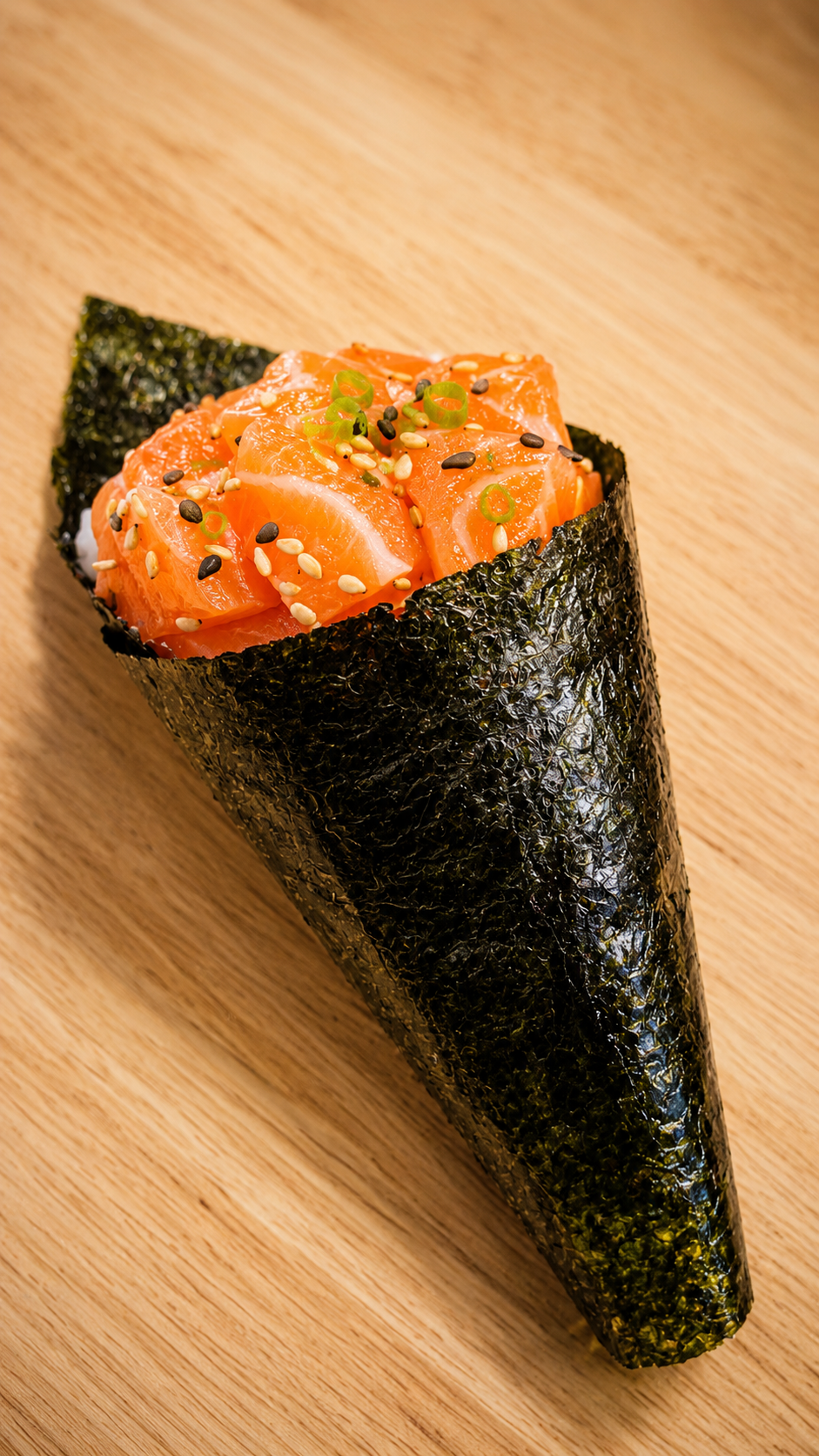 A single cone of sushi wrapped in dark seaweed with orange salmon filling, topped with sesame seeds and green onions, set on a light wood surface.