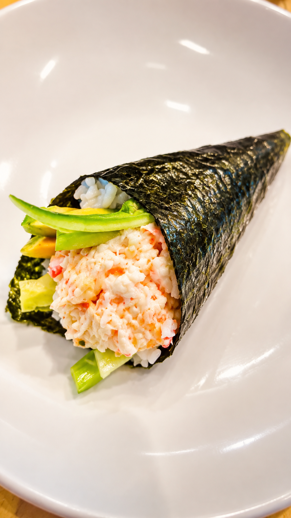 A close-up of a sushi cone filled with creamy crab meat, green avocado, and rice, wrapped in dark seaweed and served on a white plate.