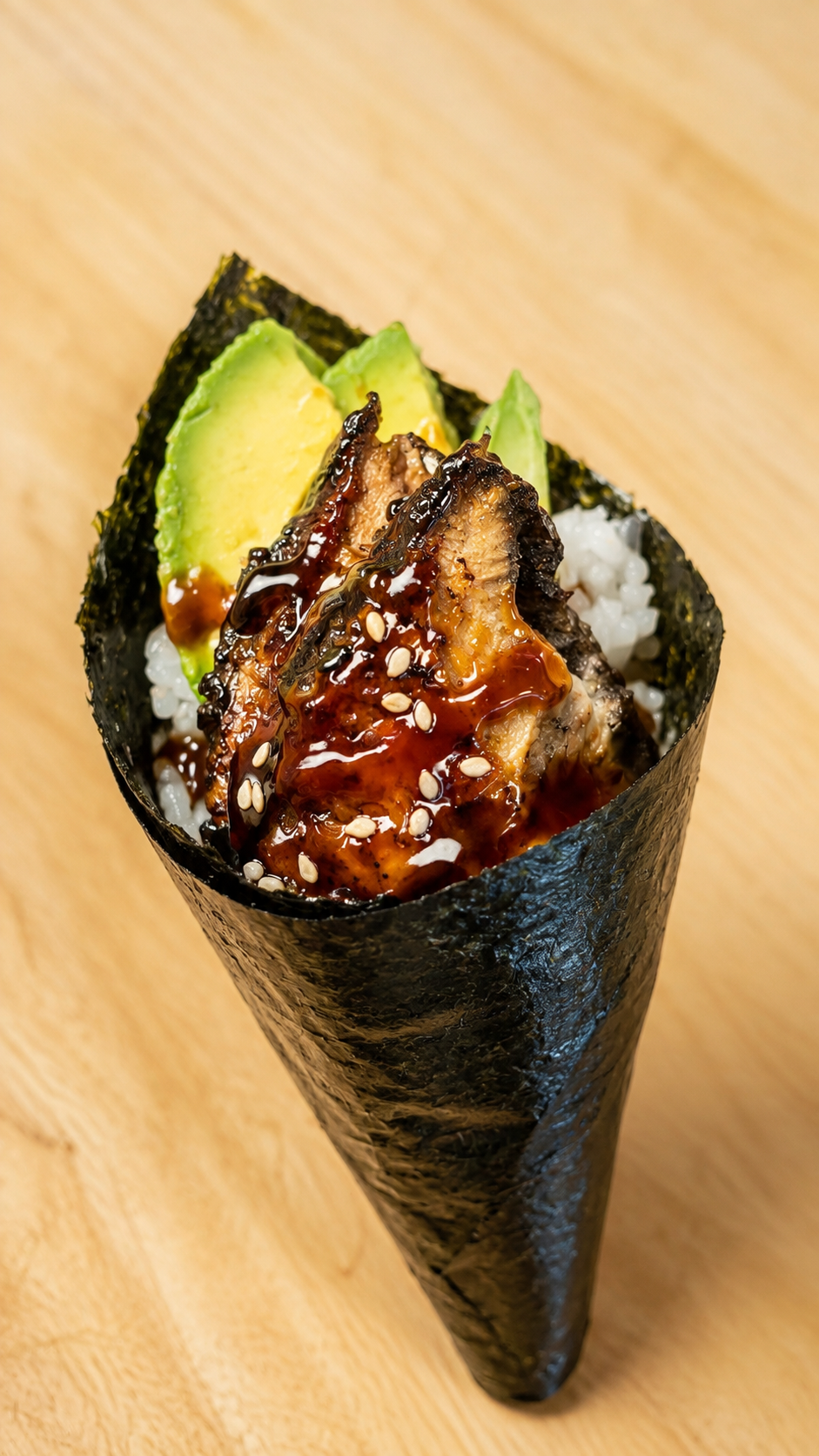 A close-up shot of an eel sushi roll wrapped in a dark nori seaweed cone, filled with white rice, sliced avocado, and glazed with a glossy dark sauce and sprinkled with sesame seeds, set on a light wood background.