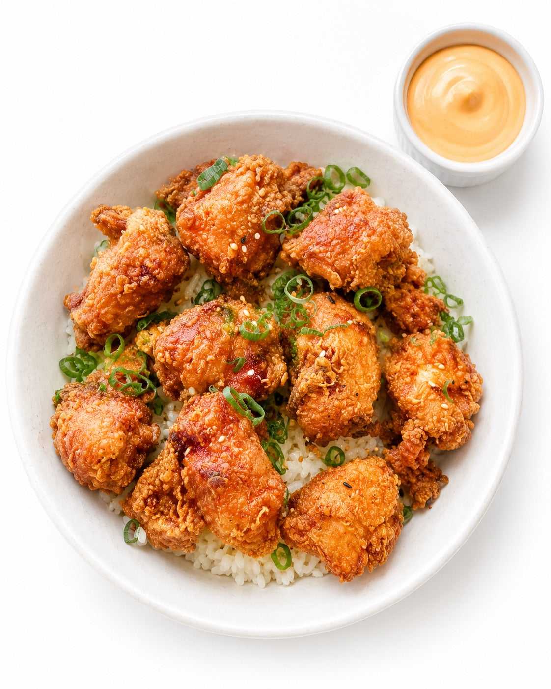 Crispy fried chicken pieces served over white rice in a white bowl, garnished with sliced green onions, with a small side dish of orange dipping sauce on a white background.