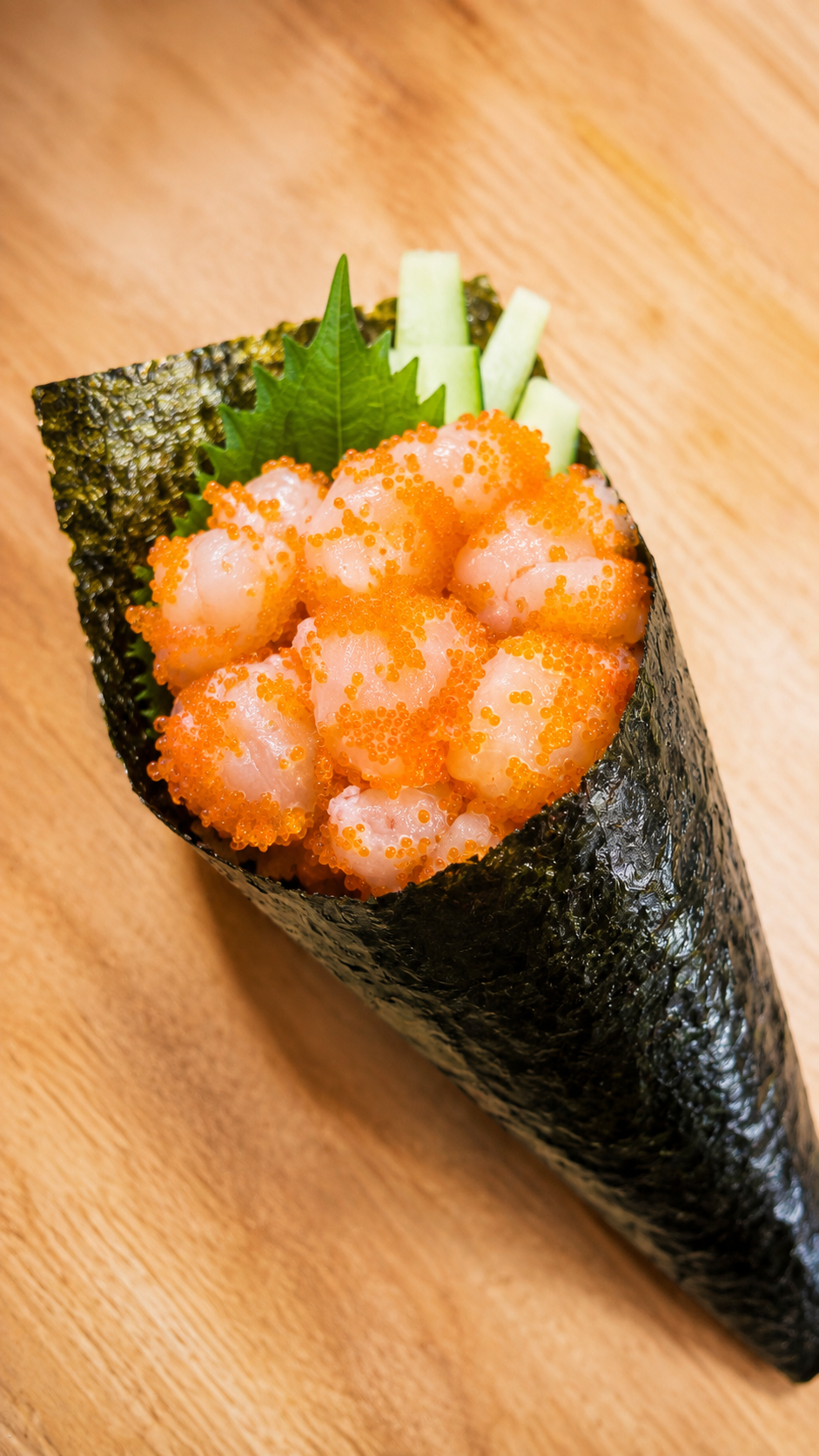 A close-up of a hand-rolled sushi cone filled with orange roe, pale pink fish pieces, cucumber sticks, and a green leaf on a wooden surface.