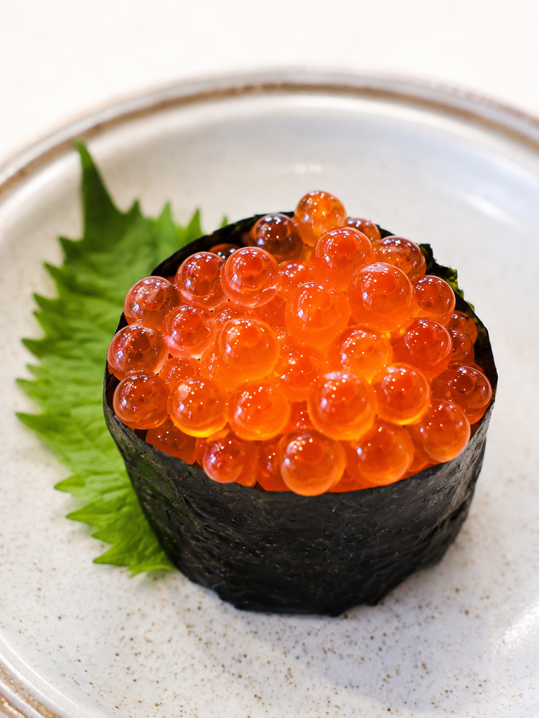 A close-up shot of a maki sushi roll topped with bright orange-red salmon roe, served on a speckled white plate with a green shiso leaf.