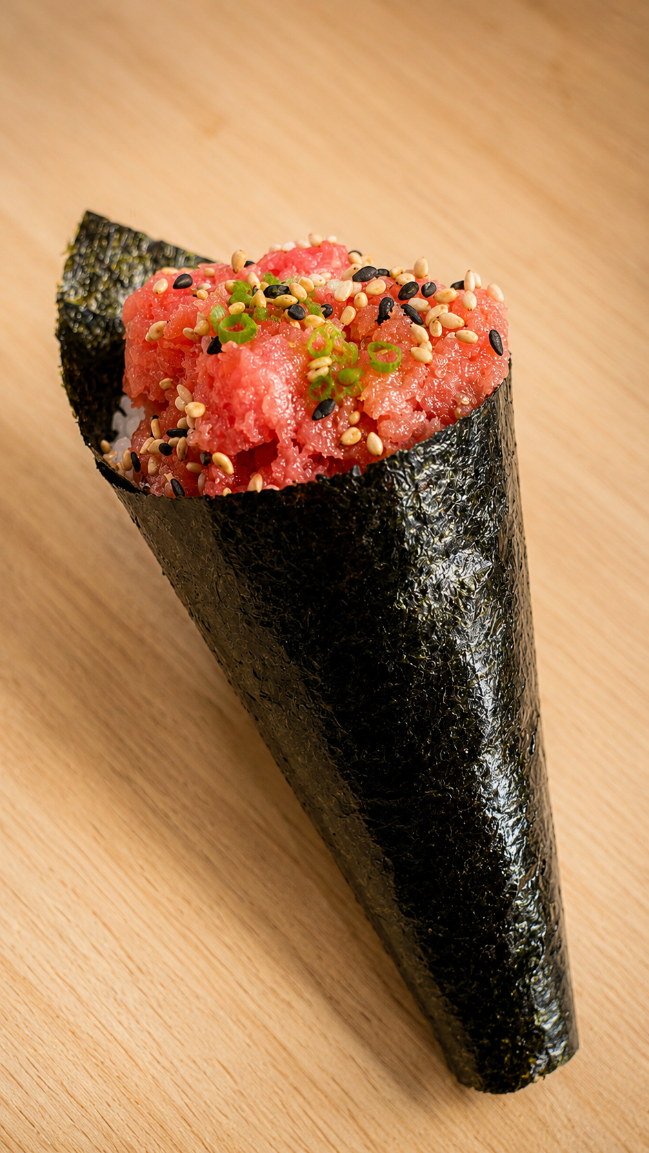 A single cone of sushi wrapped in dark seaweed with pink tuna filling, topped with sesame seeds, white sesame seeds, and chopped green onions, set on a light wood background.