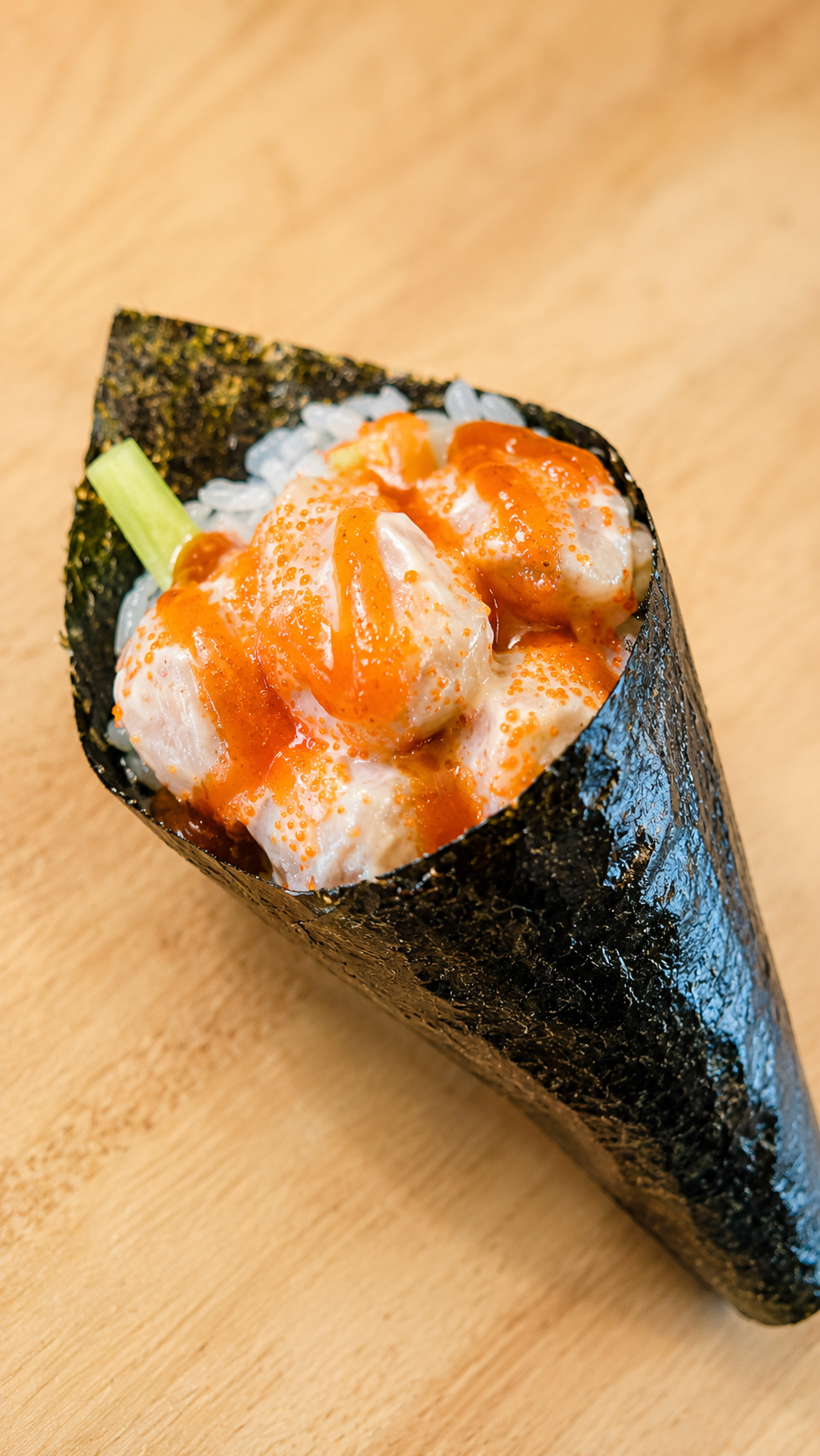 A close-up shot of a single piece of sushi on a light wood background. The sushi consists of white rice wrapped in dark seaweed, topped with a creamy white sauce and a drizzle of orange-red sauce, with a small green vegetable piece visible on the left.
